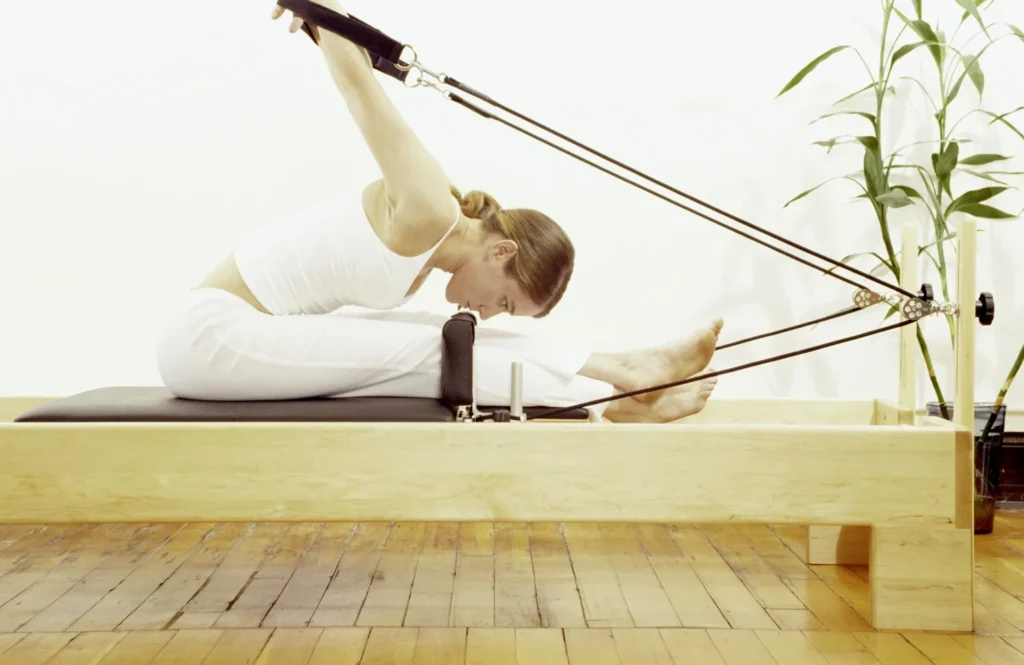 A lady with blonde hair dressed in white pants and white sleeveless top on a wooden reformer with arms behind her
